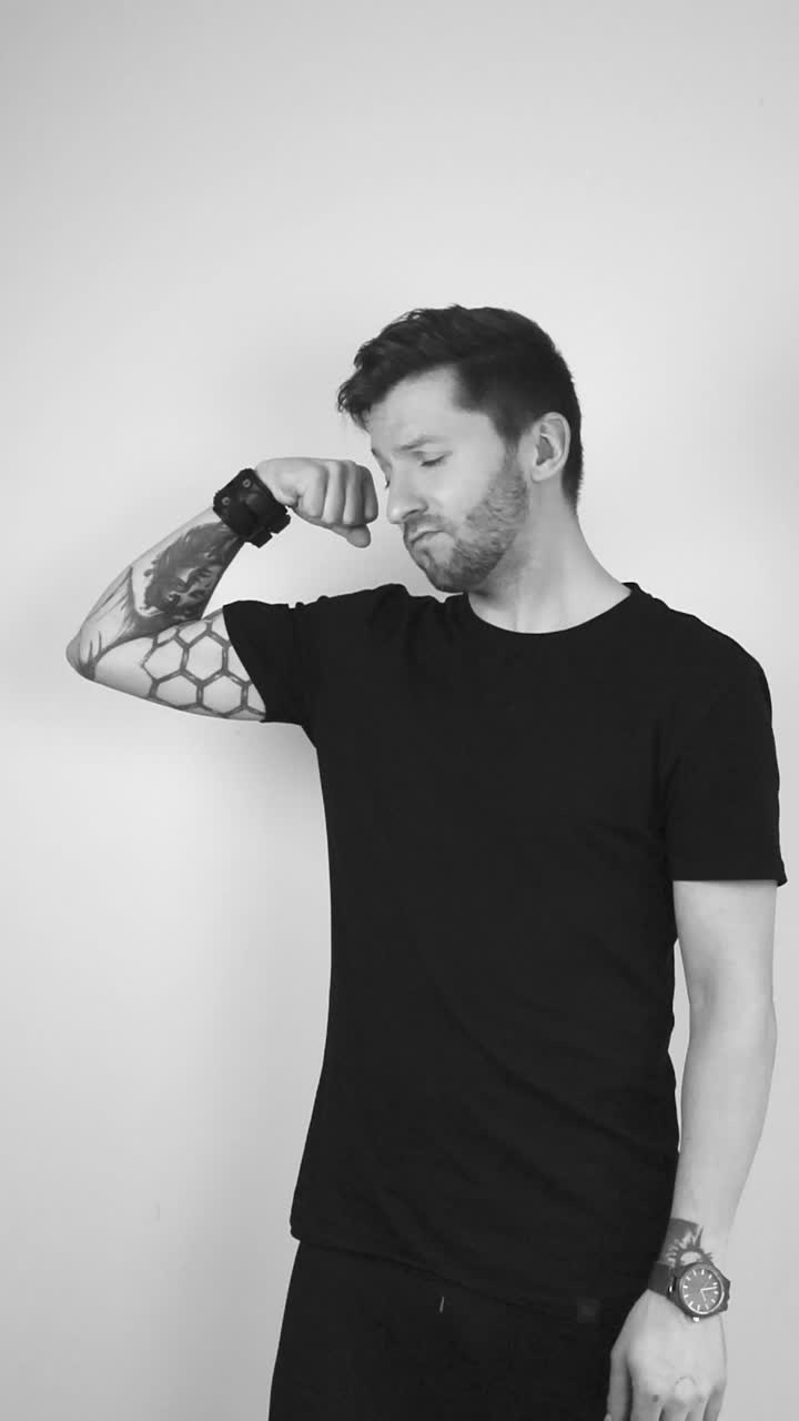 Grayscale footage of a white tattooed short haired man lifting up his arm and showing his biceps muscle and is happy. The man begins to nod after showing muscle and points at it with satisfaction.