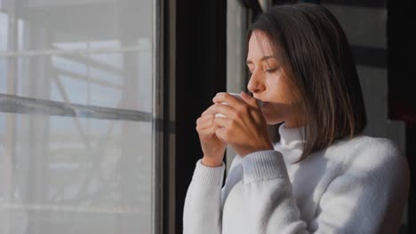 Kaukasische-Brünette-Steht-Im-Sonnenlicht-In-Der-Nähe-Eines-Breiten-Fensters-Und-Trinkt-Kaffee