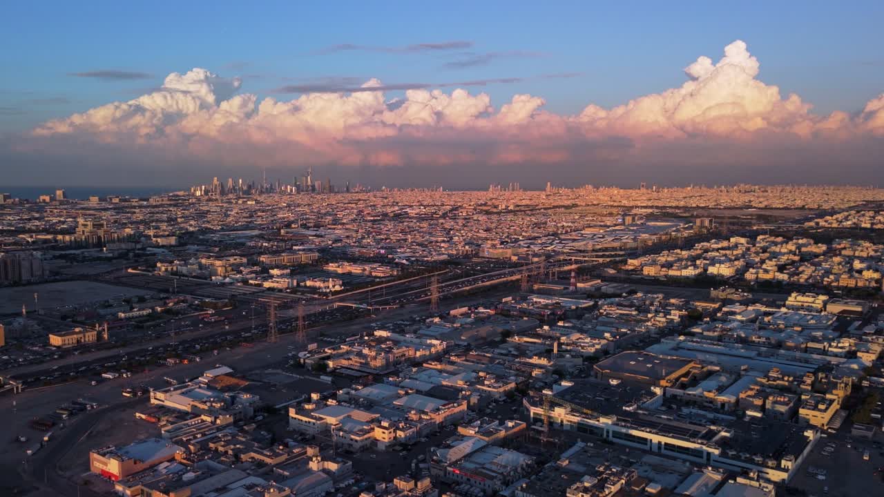 Wide drone view of Kuwait City at golden hour: distant skyline on the Gulf coast, towering cumulonimbus, vast residential grid, highways and power pylons; warm sunset glow and clear visibility