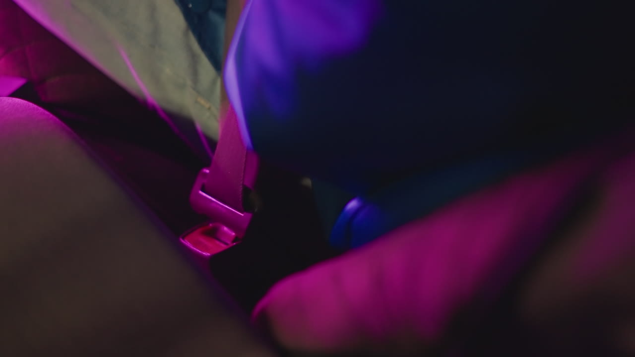 sedile posteriore al neon, pendolare che allaccia la cintura, mani ferme tirano la cintura sopra la giacca con dettagli a righe sotto una luce magenta e indaco, un primo piano stabile mostra la routine di sicurezza e l'umore tranquillo del viaggio urbano