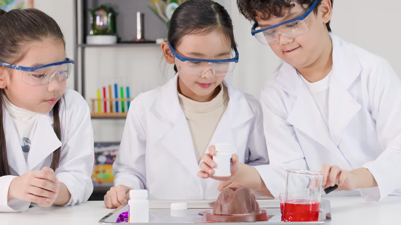 Siswa-siswi Asia yang mengenakan jas lab dan kacamata pengaman sedang melakukan eksperimen gunung berapi kimia di kelas.