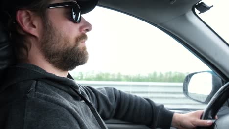 bearded man in a cap and sunglasses driving a car during road trip. side view from passenger inside car interior. summer day. male hand on steering wheel. driver staring intently at the road.