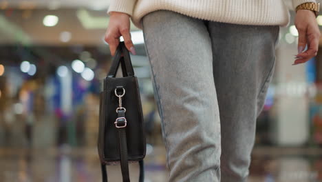 low angle close up of stylist lady carrying designer black bag walking inside well lit mall corridor on glossy tiled floor under bright ceiling lights capturing motion and fashion style