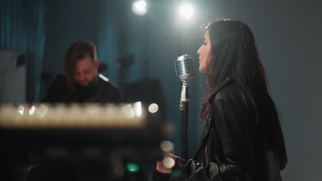 Blurry view of a singer and a guitarist, both wearing black, sitting and performing in a dimly lit room. The singer is positioned near a vintage microphone, with soft lighting