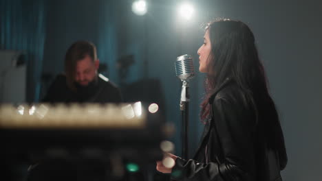 blurry view of a singer and a guitarist, both wearing black, sitting and performing in a dimly lit room. the singer is positioned near a vintage microphone, with soft lighting