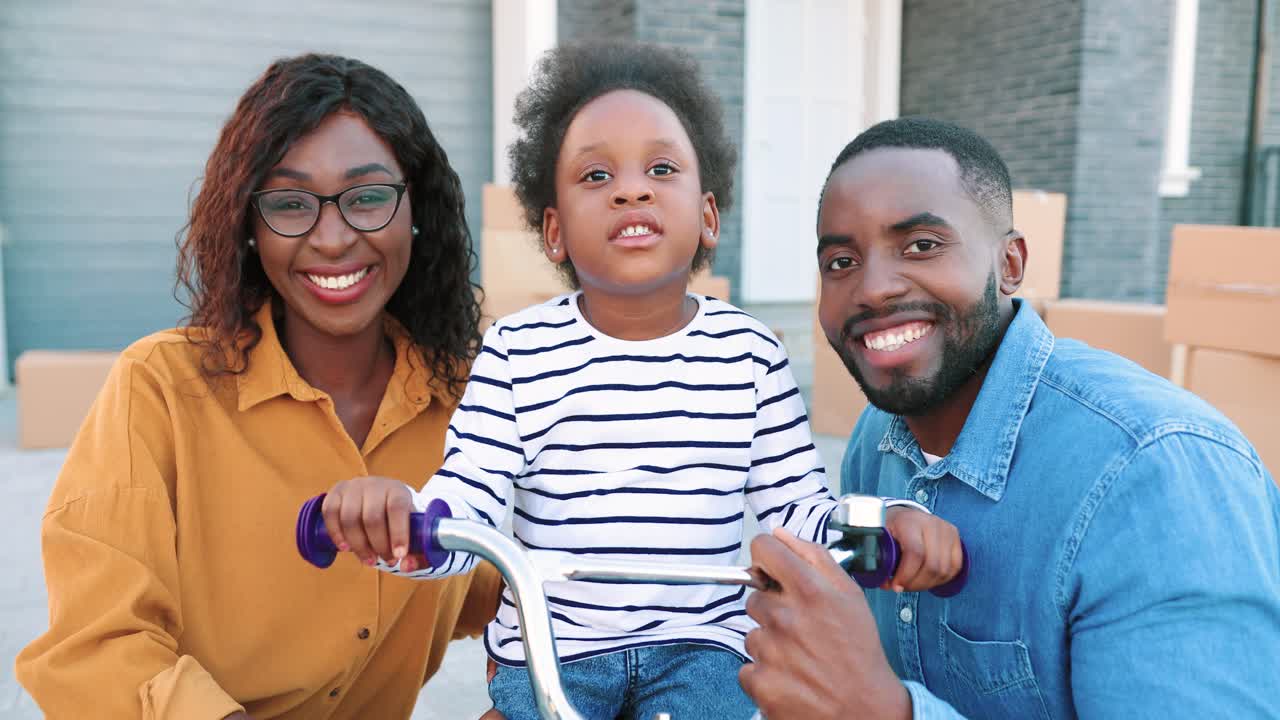 Portrait of young African American happy family with small cute girl on bike smiling to camera outdoors