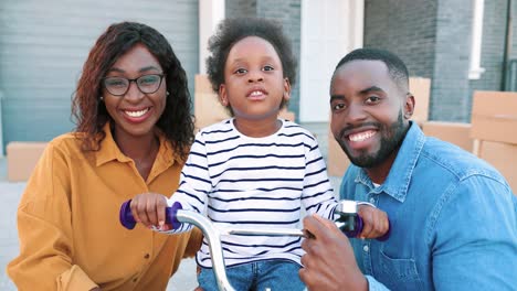 retrato de una joven familia afroamericana feliz con una pequeña y linda niña en bicicleta sonriendo a la cámara al aire libre