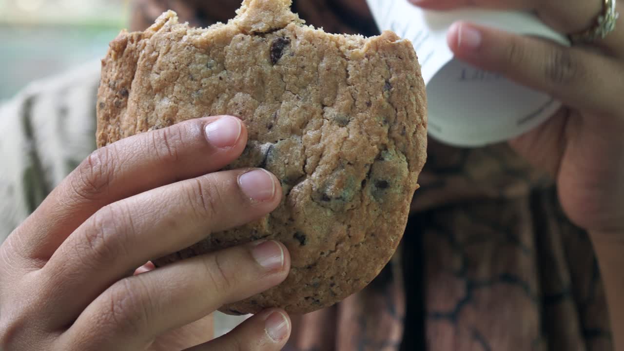 Person eating a chocolate chip cookie and drinking coffee/tea outside