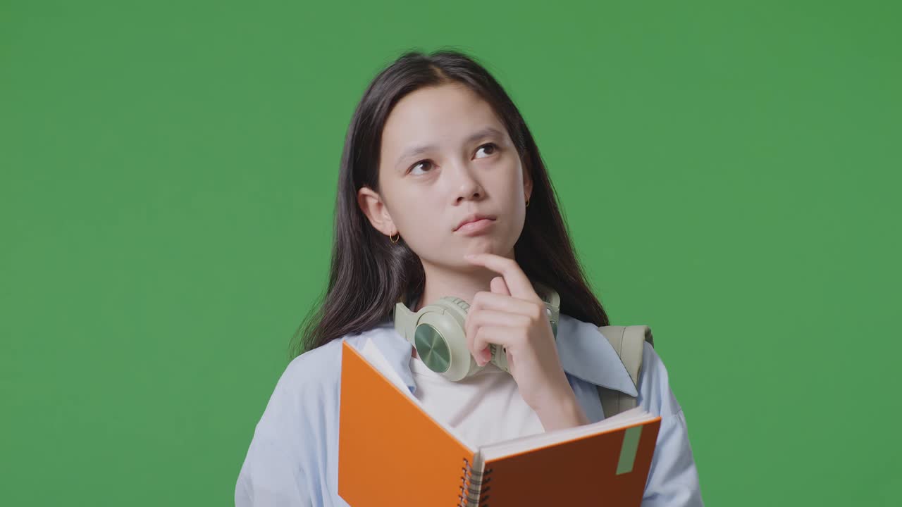 close up de una estudiante adolescente asiática con una mochila leyendo un libro y pensando y mirando a su alrededor mientras estaba de pie en el estudio de fondo de la pantalla verde