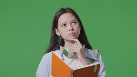 close up of asian teen girl student with a backpack reading book and thinking and looking around while standing in the green screen background studio