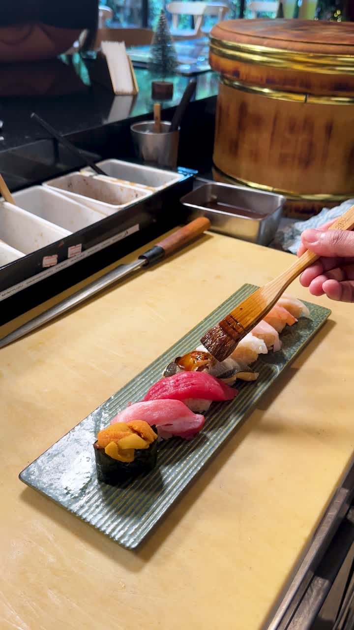 Close-up de um chef de sushi pincelando uma travessa de nigiri variado com molho num balcão tradicional.