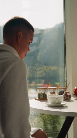 joven mueve la silla a la mesa por la mañana. el marido viene a la cocina para tener un desayuno saludable por la ventana con vista a la naturaleza. rituales matutinos durante el tiempo de descanso