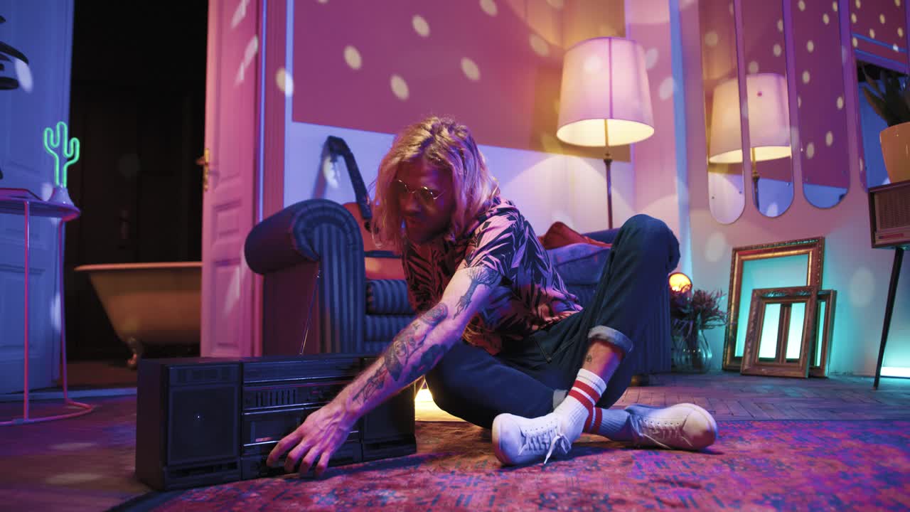 feliz joven caucásico con gafas de sol sentado en el suelo, encendiendo la caja de resonancia y escuchando su música favorita en una sala de estar vintage