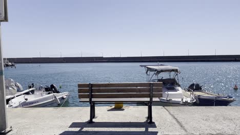 A wooden bench overlooks a calm harbor with docked boats, clear blue water, and a distant stone breakwater under a bright sky
