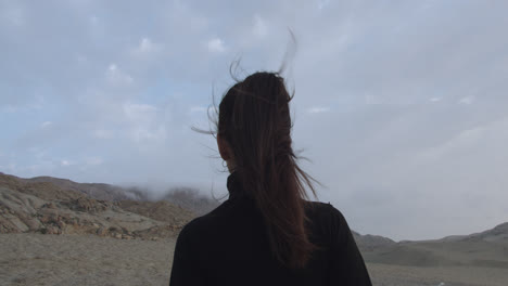 primer plano de la espalda de una mujer misteriosa vestida de negro de pie mirando el desierto con un cielo nublado por la tarde