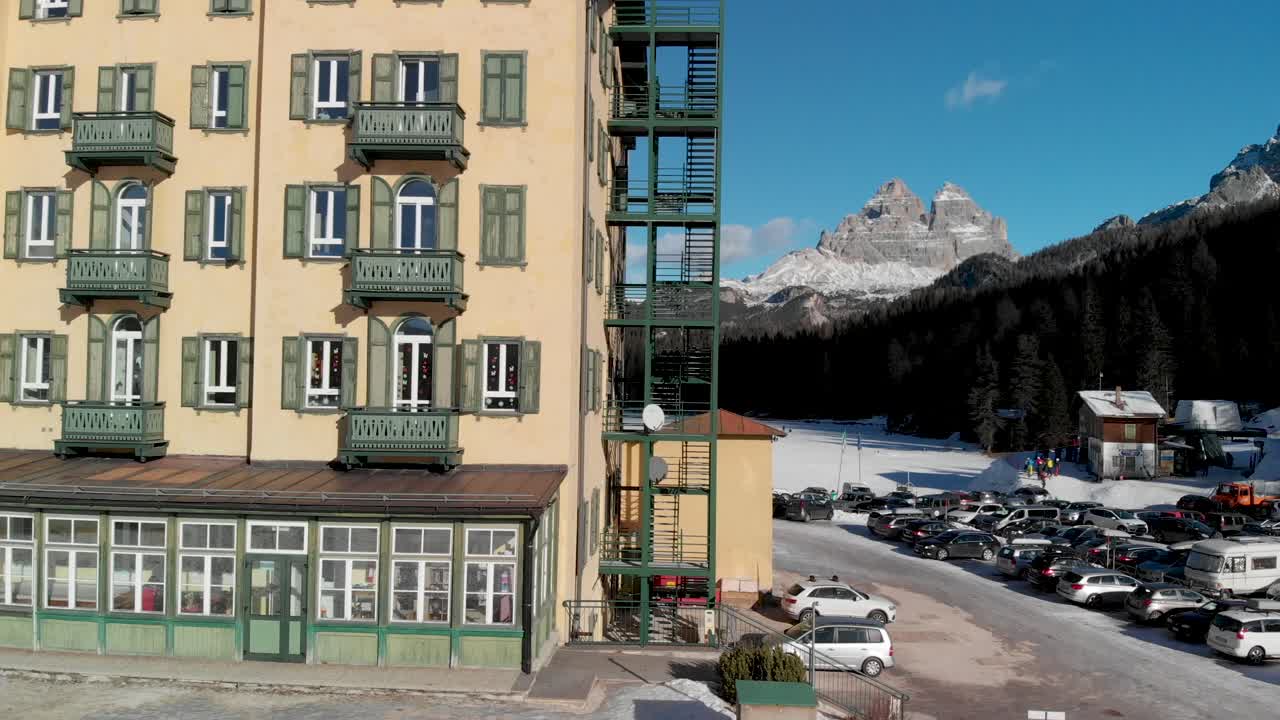 toma aérea de pedestal descendente de un gran edificio con un lago misurina congelado y los tres picos italianos de lavaredo en la distancia