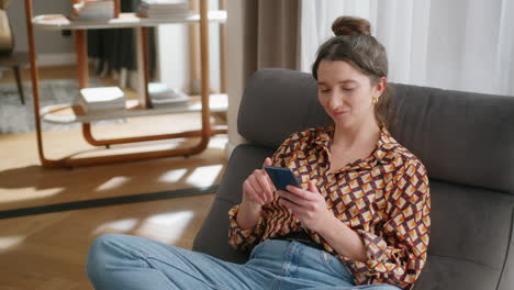 Smiling-young-woman-sits-in-living-room-and-scrolls-on-phone,-handheld