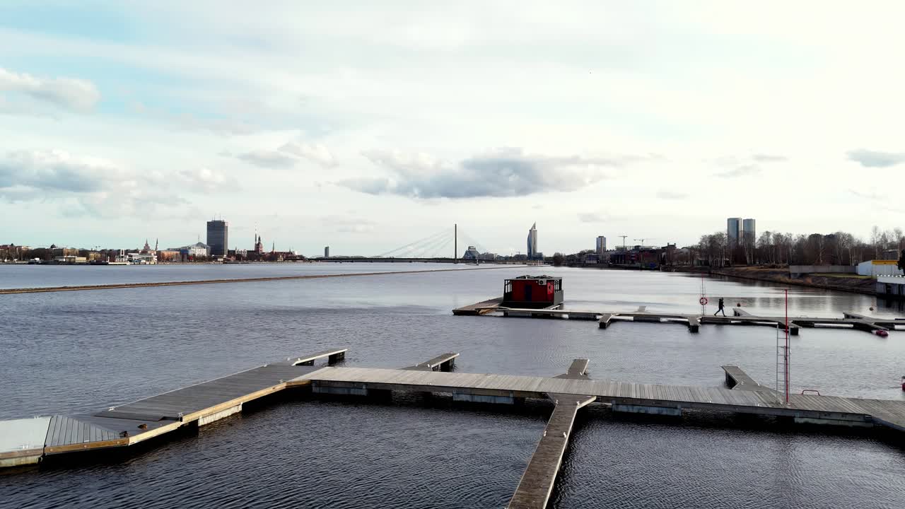 A long, wooden dock juts out into a large, calm body of water , river Daugava on a sunny day, calm body of water with a few boats docked at the marina