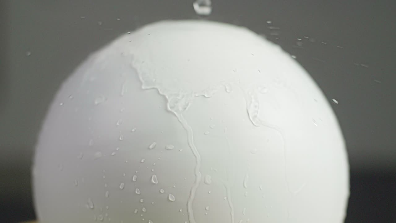 Close up of a white plain sphere wet with water running down the sphere and reflecting water drops on the sphere. A dark gray background. Shot in Slow Motion in 4K.