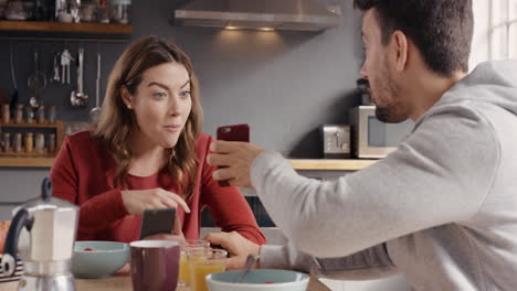 excited couple using smart phone devices eating breakfast at home sharing social media news from app