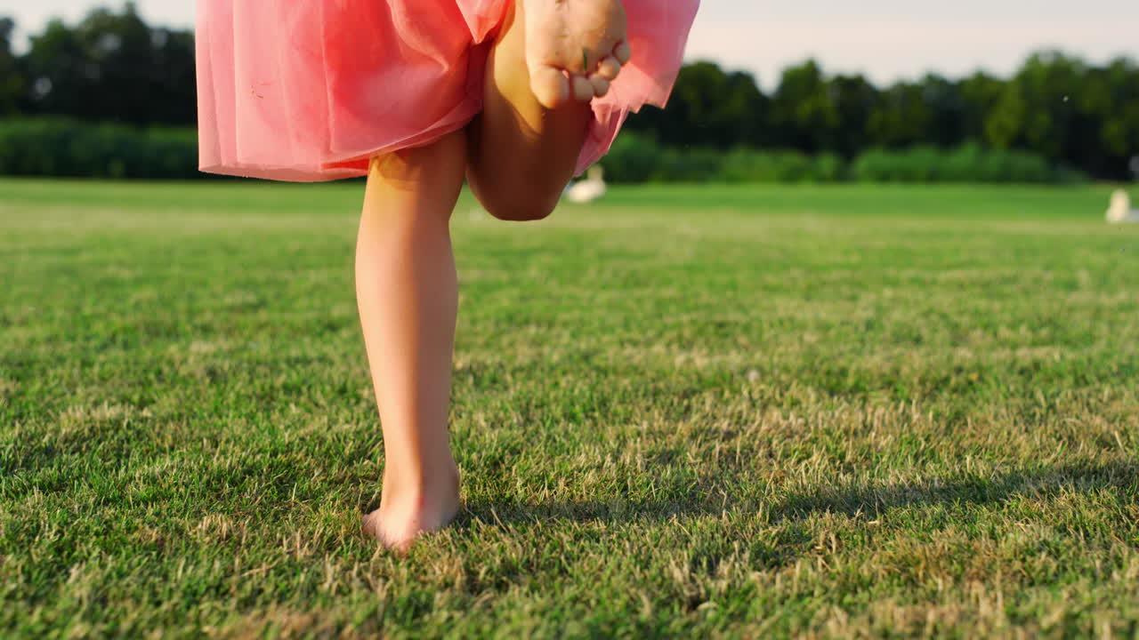Rear view of unrecognizable girl running barefoot on green grass at city park.