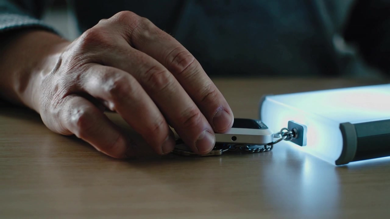 Hand entering, lowering white device near blue fob on wood table, aligning with lit port, charging
