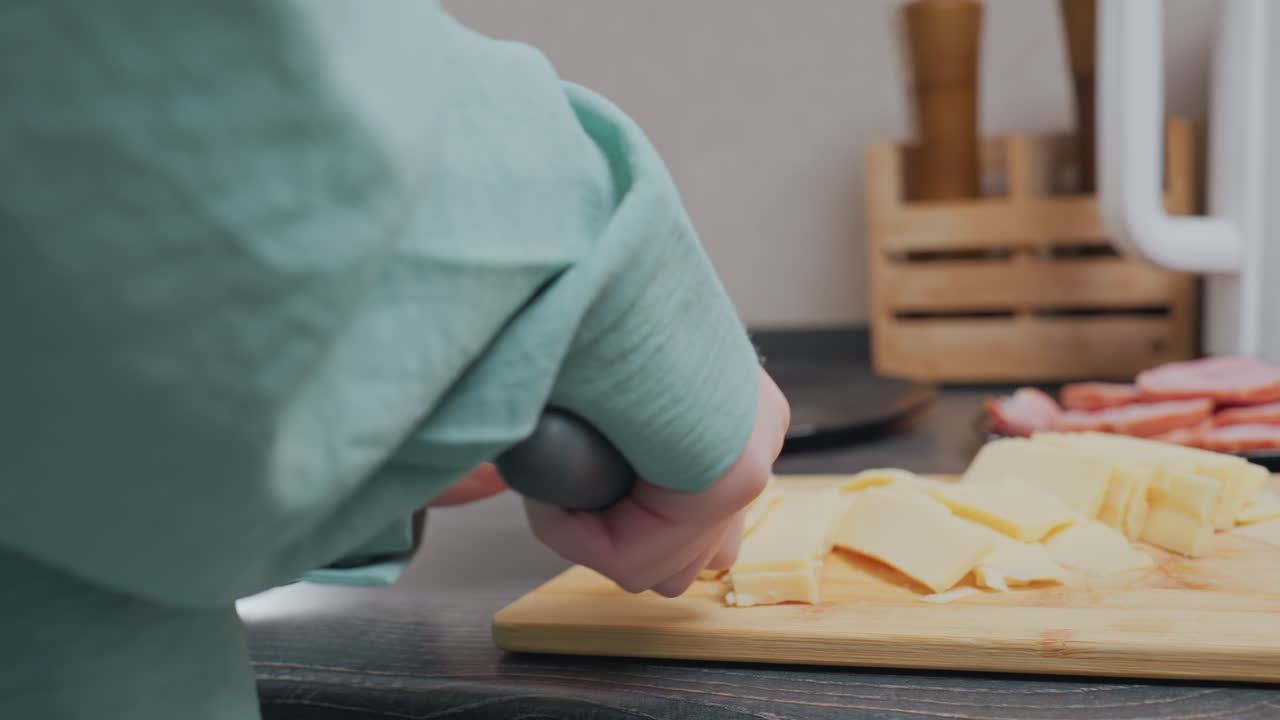 rear view of woman in green shirt slicing yellow cheese on cutting board in kitchen with sliced meat on plate, electric kettle, and spice jars in wooden holder on countertop in soft natural lighting