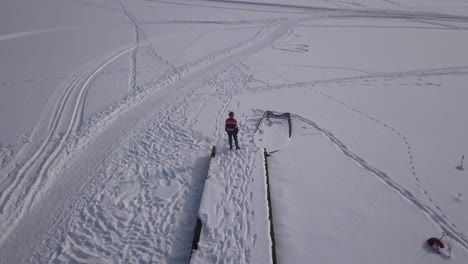 Slow-panning-aerial-footage-of-blonde-female-standing-on-edge-of-wooden-jetty-staring-over-frozen-lake