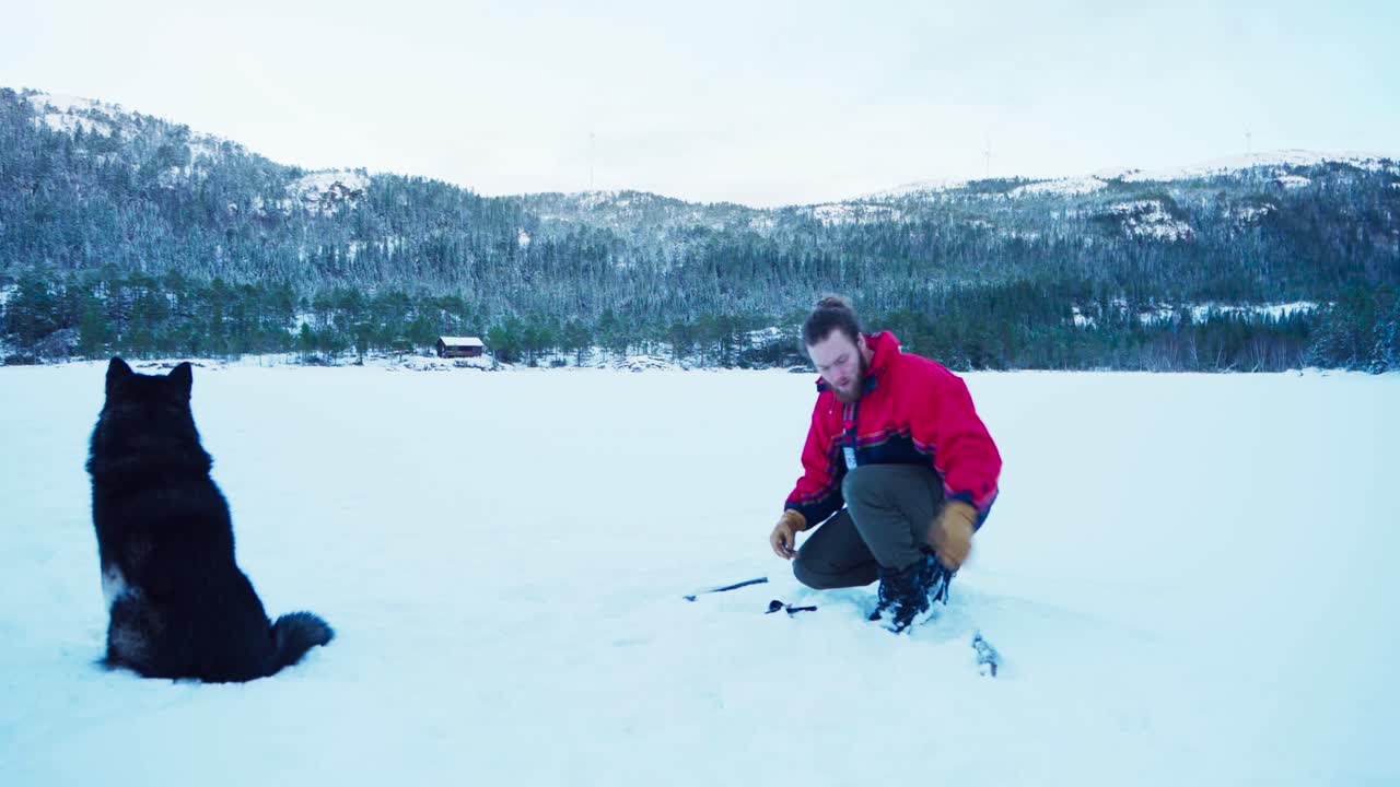 Uomo e il suo cane seduti sulla neve, che pescano nel ghiaccio in Norvegia in inverno