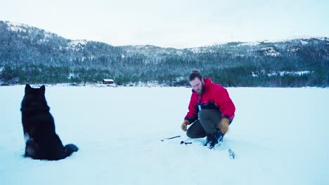 Hombre-Y-Su-Perro-Mascota-Sentados-En-La-Nieve,-Pescando-En-Hielo-En-Noruega-Durante-El-Invierno