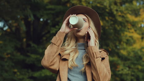 Close-up-view-of-caucasian-young-blonde-woman-wearing-a-hat-and-talking-on-the-phone-and-drinking-coffee-to-go-in-the-park-in-autumn