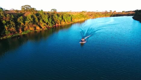 Ein-Kleines-Fischerboot-Fährt-In-Der-Abendsonne-Einen-Tiefblauen-Fluss-Hinunter,-Drohnenaufnahme-Aus-Der-Luft