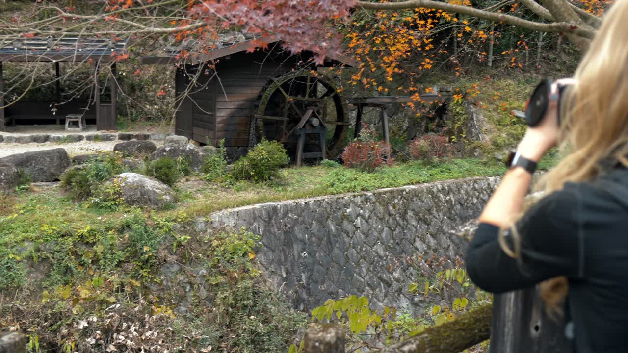 A photographer captures the stunning beauty of a traditional water mill along the historic Nakasendo Trail in Japan, surrounded by vibrant red autumn foliage.