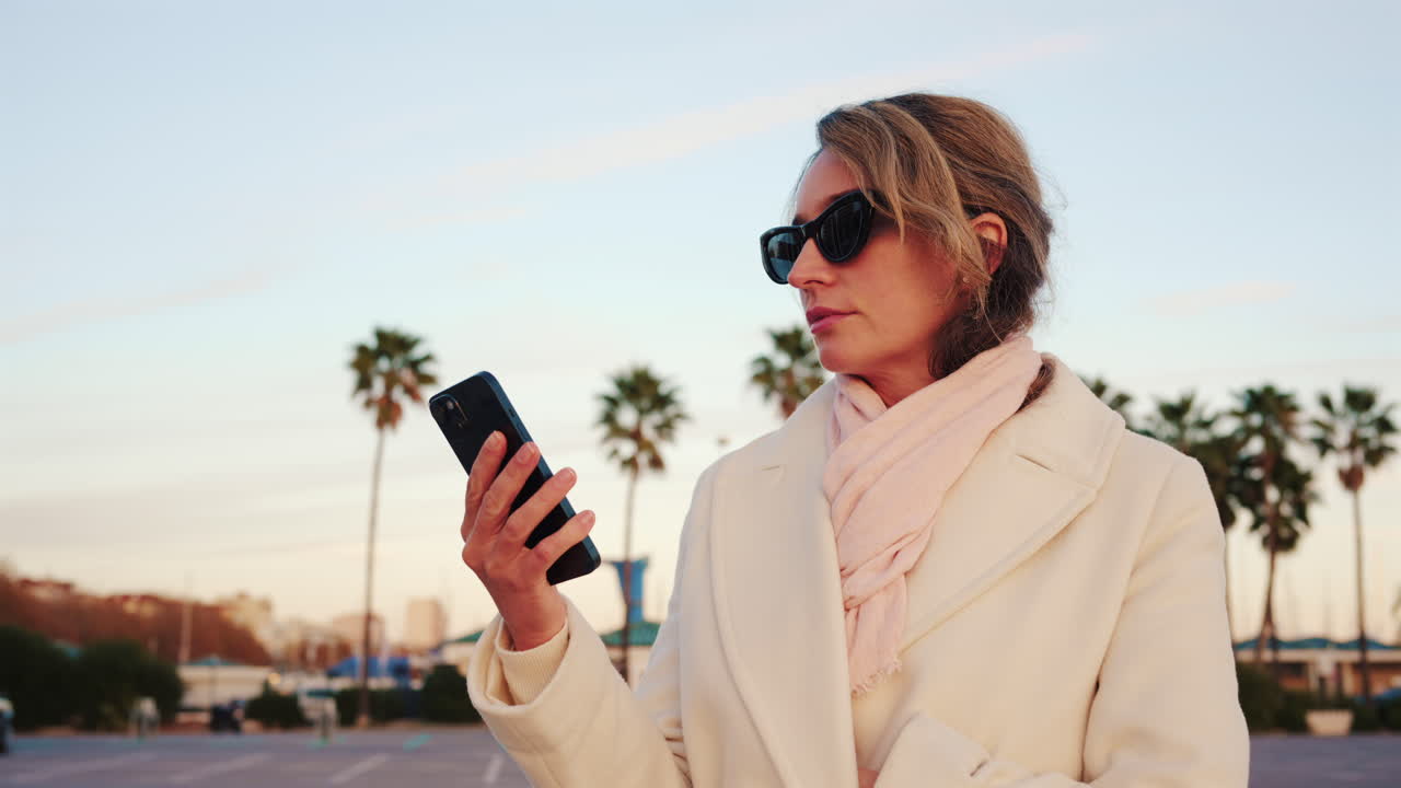 Donna elegante con cappotto bianco, sciarpa e occhiali da sole che controlla il telefono in una zona esterna soleggiata con palme