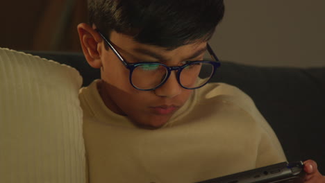 young boy sitting on sofa at home playing game or streaming onto handheld gaming device at night