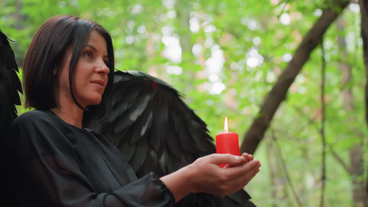 Side view of black fairy with dark wings holding glowing red candle while standing in green forest, smiling softly during tranquil moment, symbolizing mystery, light, faith, and harmony with nature