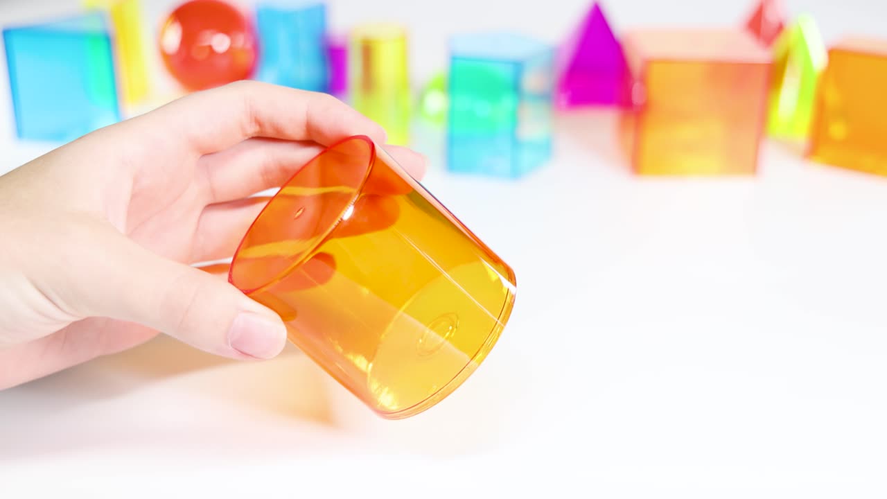 Hands manipulate vibrant 3D geometric shapes on a white surface, highlighting educational and playful exploration in bright lighting