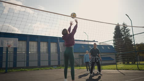 coach training beginner in volleyball, guiding movements while two female players observe in an outdoor court, volleyball net, basketball hoop, and sports facility with a blue building in background