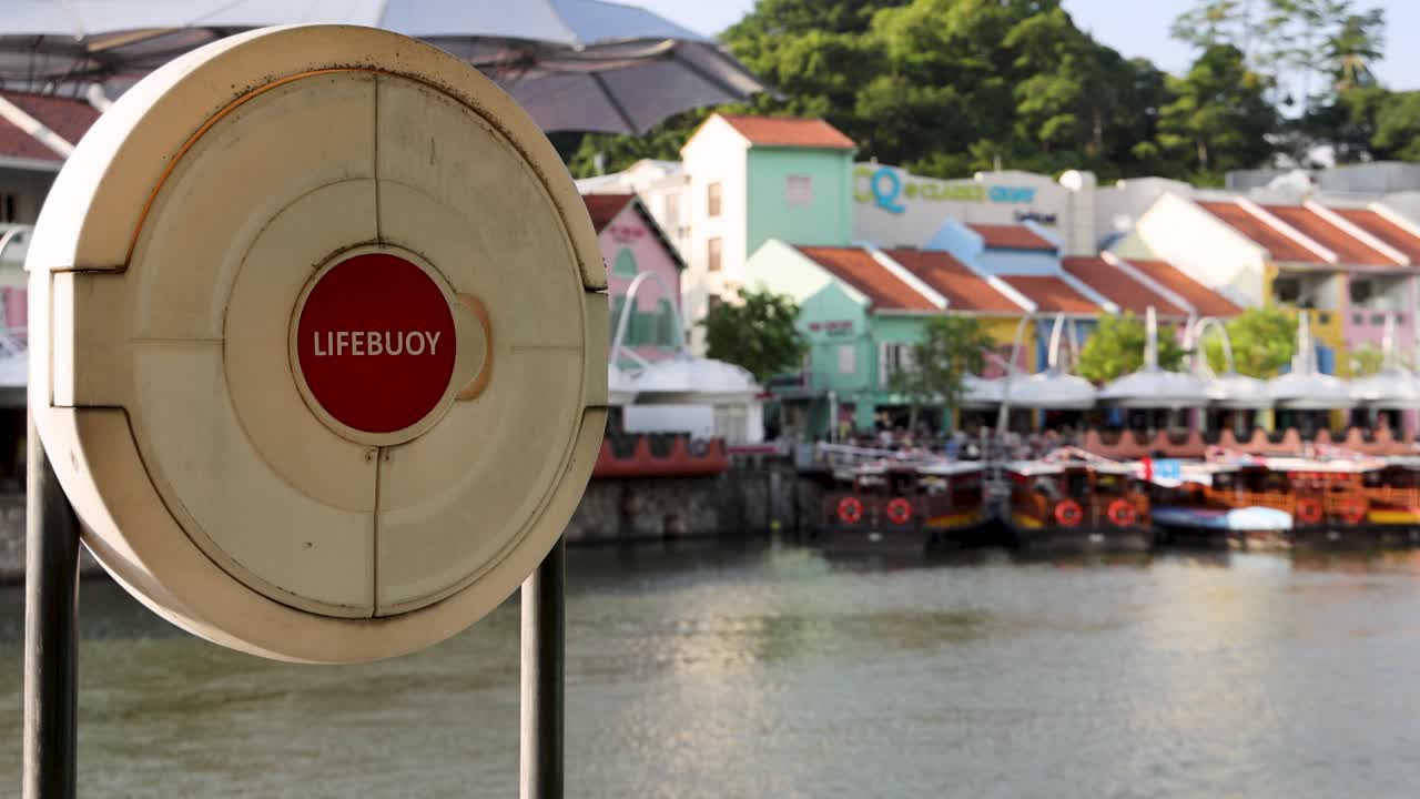 A stationary lifebuoy is prominently displayed in the foreground as the camera slowly pans to reveal colorful riverside shophouses along Singapore’s Clarke Quay in bright daylight