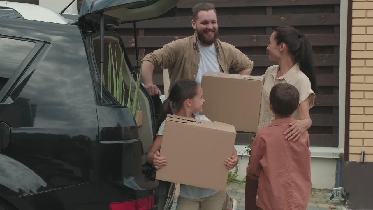 Family With Two Children Taking Boxes And Personal Items From Car For Moving