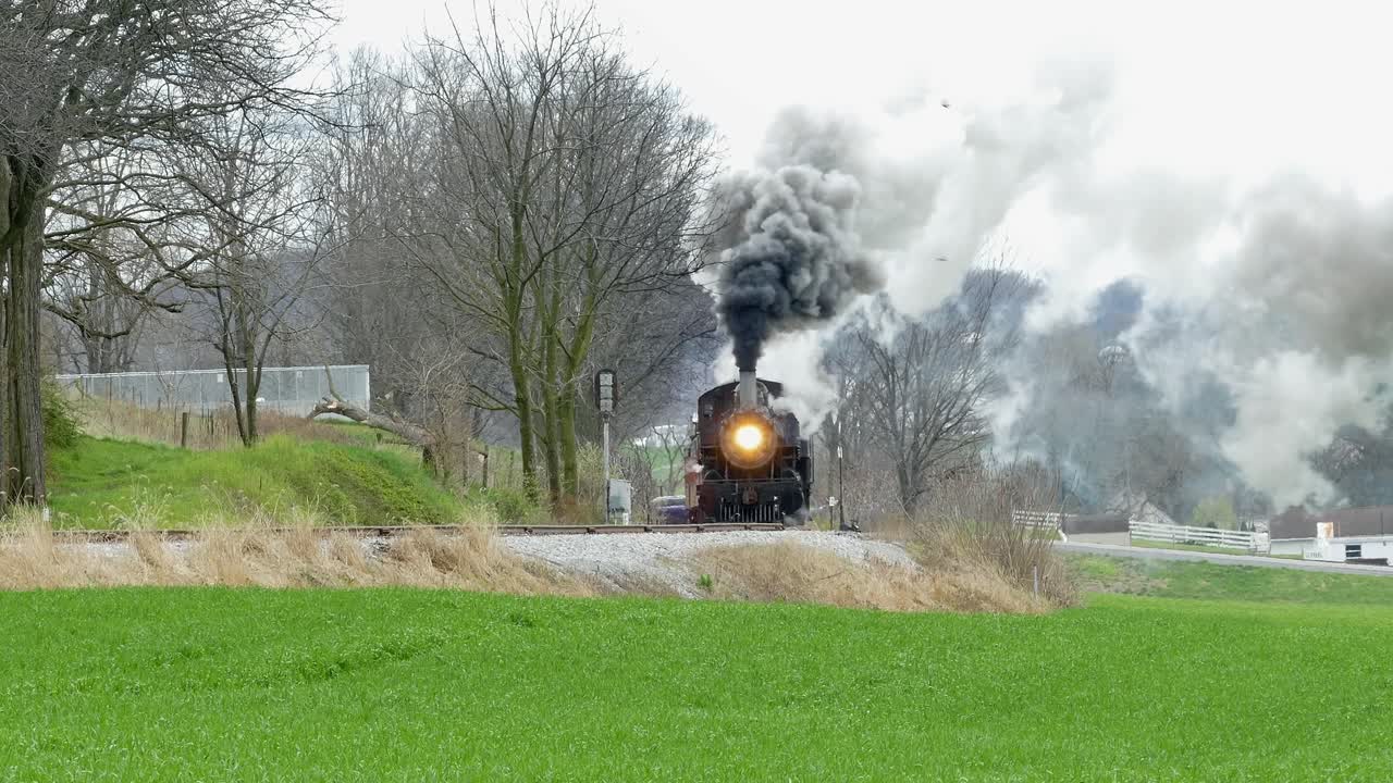 A classic steam train moves along the tracks surrounded by lush green fields and trees. The day is bright with fluffy clouds, creating a perfect setting for train enthusiasts and nature lovers