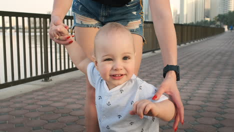 un niño sonriente sosteniendo la mano de su madre hace los primeros pasos caminando por el paseo marítimo en verano.