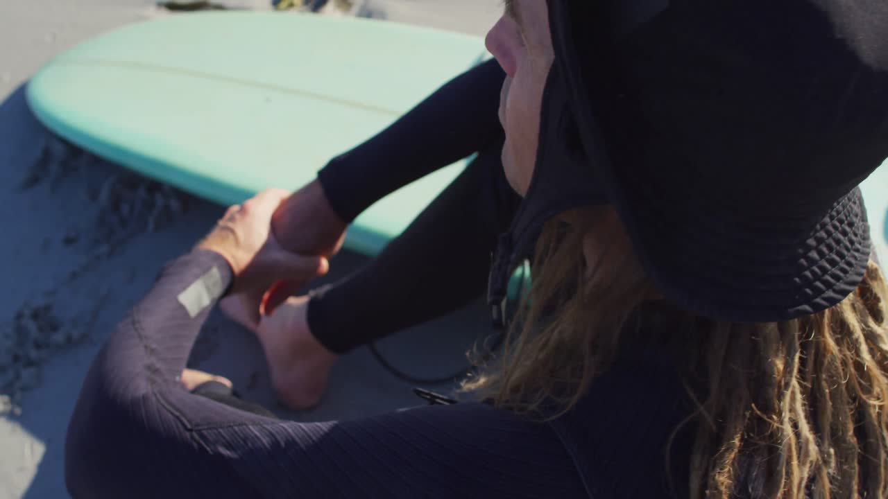 Video of smiling caucasian man with dreadlocks in wetsuit sitting on sunny beach with surfboard