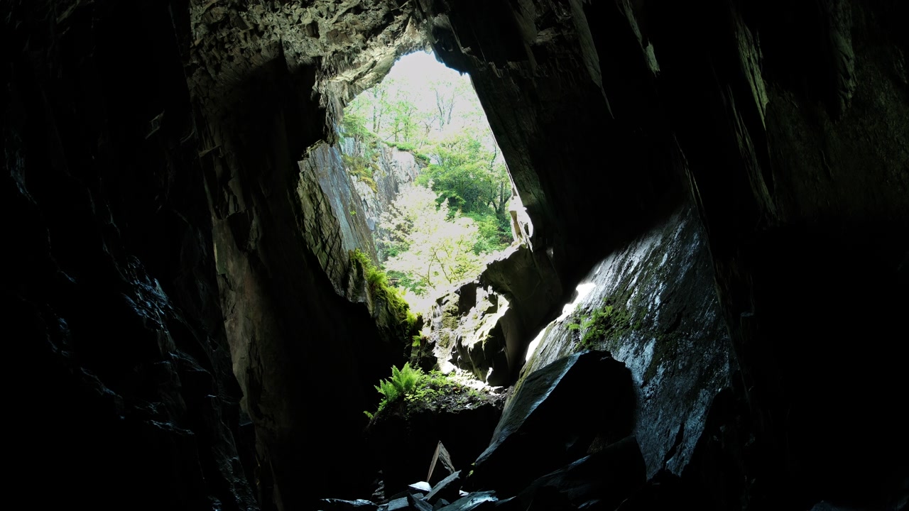 Ripresa statica dall'interno della Cathedral Cavern a Little Langdale, che guarda verso l'ingresso illuminato dal sole della grotta, mostrando imponenti formazioni rocciose, pietra coperta di muschio e il drammatico paesaggio sotterraneo.