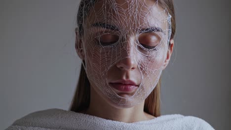 female wearing white mesh face mask relaxing during rejuvenating beauty treatment in minimalist gray studio, embodying skincare wellness and cosmetic self care