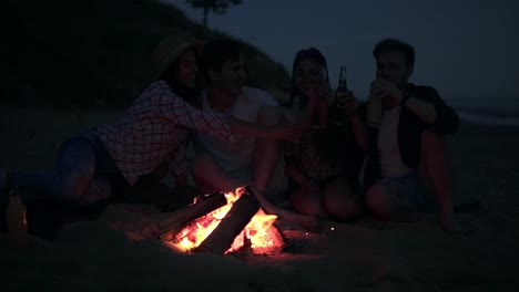 picnic of young people with bonfire on the beach in the evening drinking beer and doing cheers. happy friends singing songs and