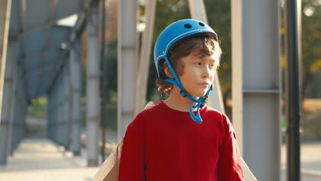 Portrait-Of-A-Cute-Little-Boy-In-Helmet-And-Red-Sweater-Looking-At-The-Camera-While-Standing-On-A-Bridge-On-A-Sunny-Day