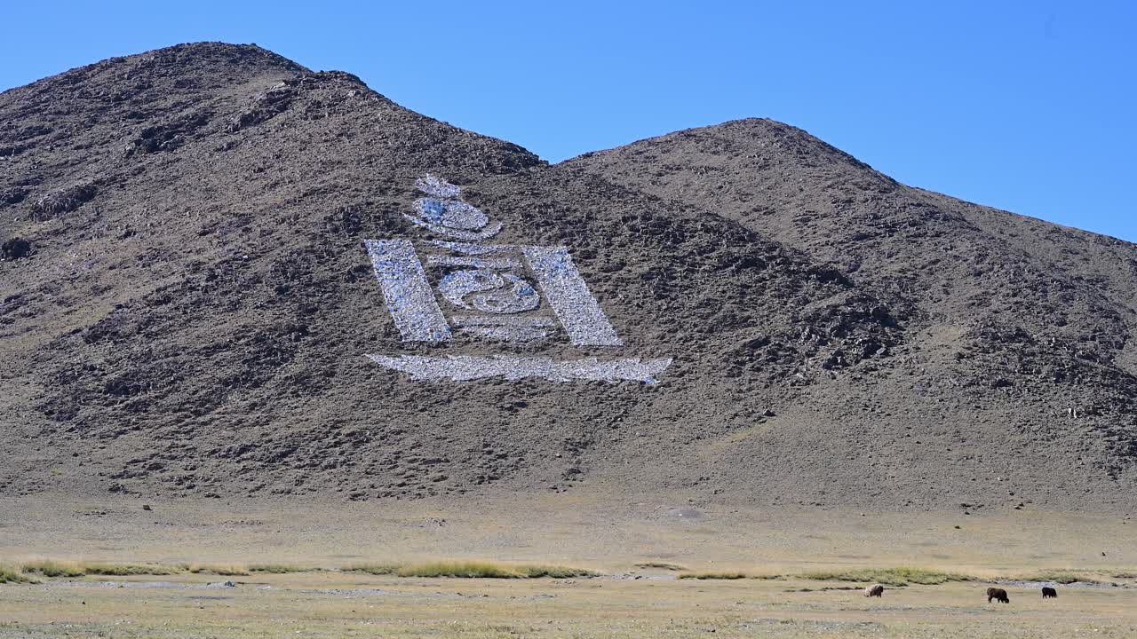 A wide shot of the Mongolian landscape reveals the national Soyombo symbol made from stones on a rocky hill. The cultural landmark stands out against the arid, remote terrain