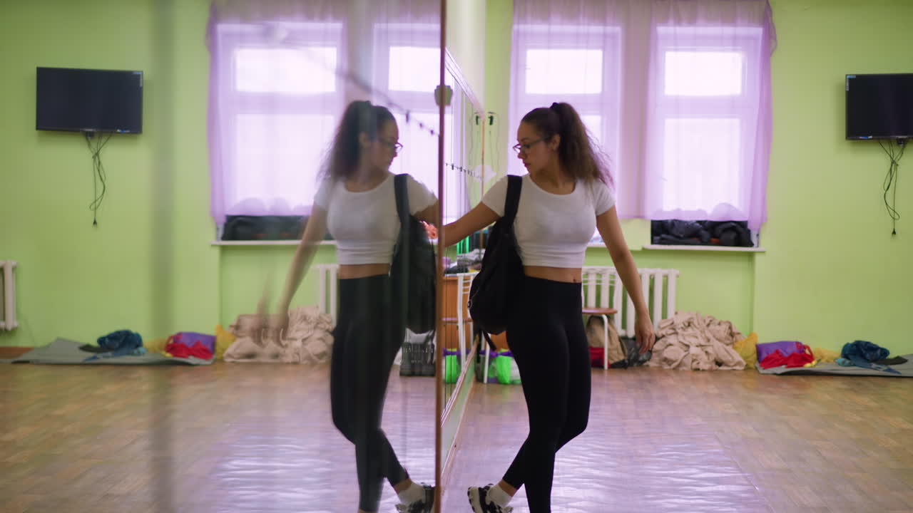 Back view of lady in casual white top and black leggings carrying school bag walking towards window, body reflected clearly in large wall mirror inside bright room with wooden floor
