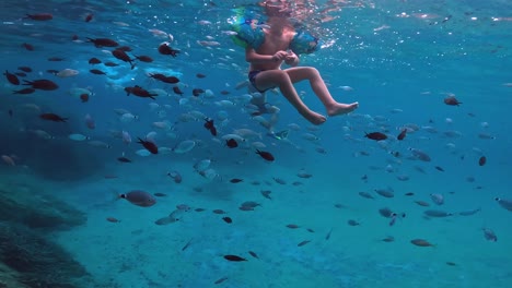 Vista-Submarina-De-Niños-Pequeños-Nadando-Con-Brazaletes-Inflables-En-Medio-De-Una-Gran-Escuela-De-Peces-En-Aguas-Azules-Profundas,-Cámara-Lenta